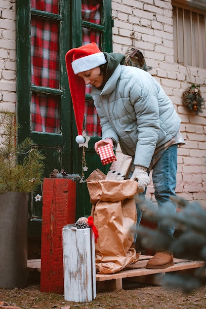 Mujer con gorro de Papá Noel lleva una bolsa de regalos desde la puerta principal del porche de la casa.