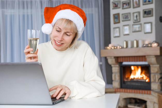 Una mujer con un gorro de Papá Noel sostiene una copa de champán, familiares y amigos en un portátil con Navidad