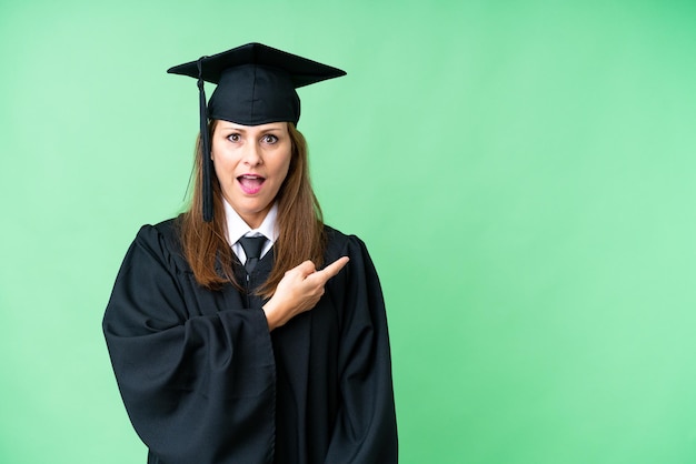 Mujer graduada universitaria de mediana edad sobre un fondo aislado sorprendida y apuntando hacia el lado