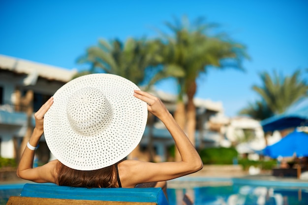 Mujer con gran sombrero blanco acostado en una tumbona cerca de la piscina del hotel, concepto de horario de verano para viajar