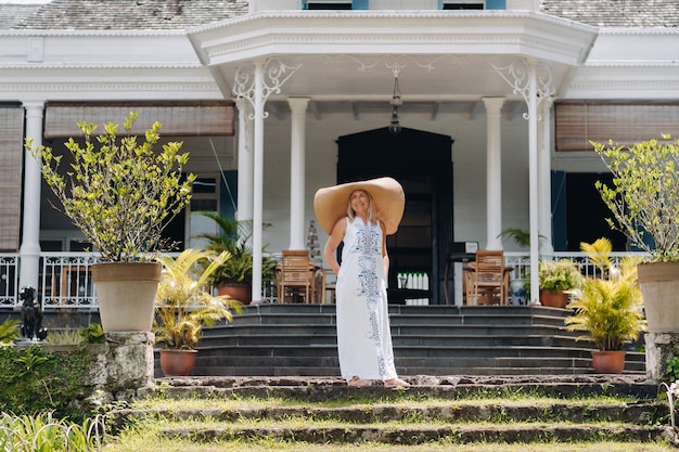 La mujer del gran sombrero sonríe una hermosa chica con un gran sombrero y un vestido blanco sonríe fuera de un antiguo edificio colonial en la isla de Mauricio