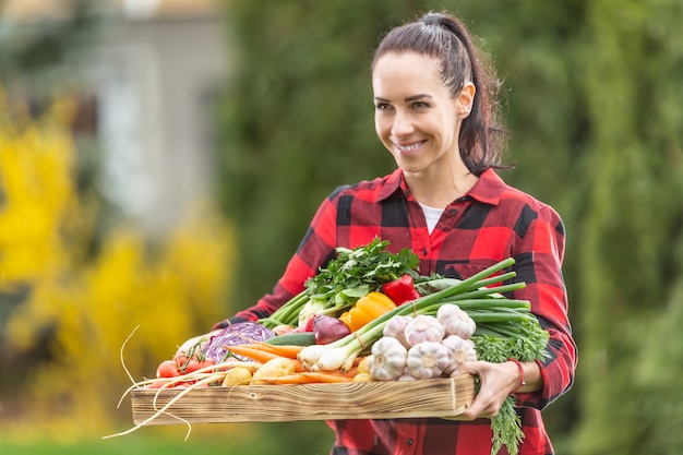 Mujer de la granja de pelo oscuro hermosa que sostiene la cesta de madera llena de verduras frescas y saludables de cosecha propia del jardín.