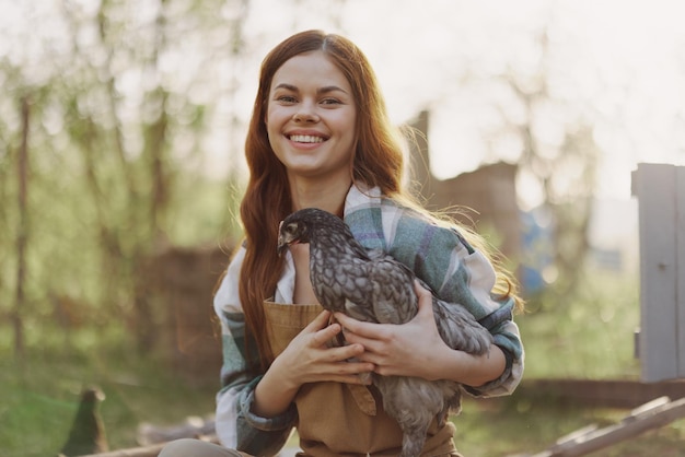 Una mujer en una granja sosteniendo un pollo y sonriendo felizmente en una granja orgánica en el atardecer de verano