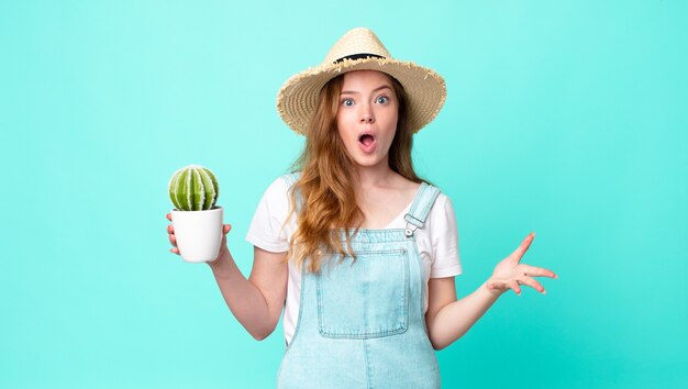 Mujer de granjero bonita cabeza roja que se siente extremadamente conmocionada y sorprendida y sosteniendo un cactus