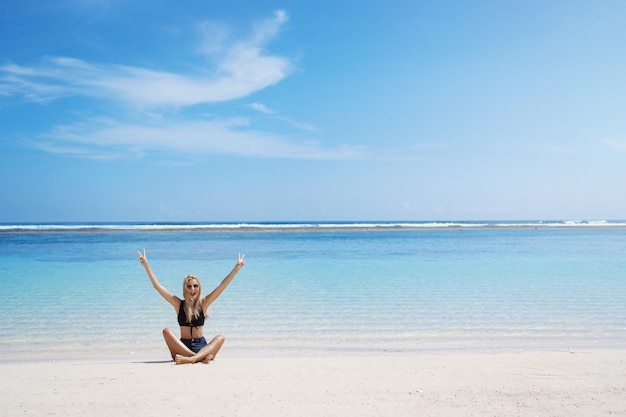 La mujer grita felizmente sentada con las piernas cruzadas, soleada playa de arena, levanta las manos, muestra la paz, el signo de la victoria, sonriendo, encantada, disfruta de las vacaciones de verano, el balneario perfecto, la gira turística, las vacaciones, las vacaciones, se relaja, está emocionado.