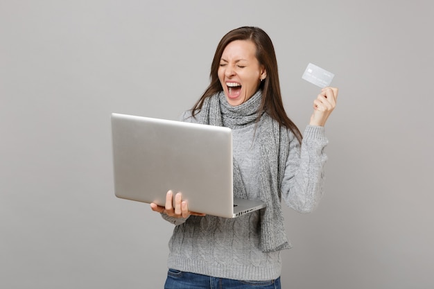 Mujer gritando en suéter, bufanda con los ojos cerrados trabajar en computadora portátil con tarjeta bancaria de crédito aislada sobre fondo gris. Estilo de vida saludable, consultoría de tratamiento en línea, concepto de estación fría.