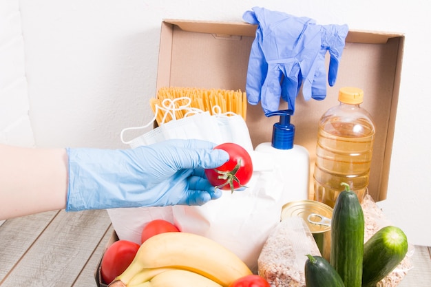 Una mujer con un guante de goma pone un tomate en una caja con alimentos y productos de higiene para donación