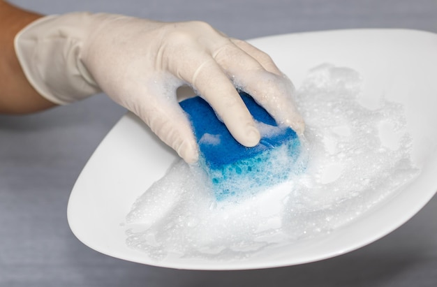 mujer con guantes blancos sosteniendo una cocina azul con plato de espuma líquida la mano está lavando un plato blanco