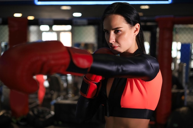 Mujer con guantes de boxeo rojos y ropa deportiva se para en un estante en la sala de boxeo