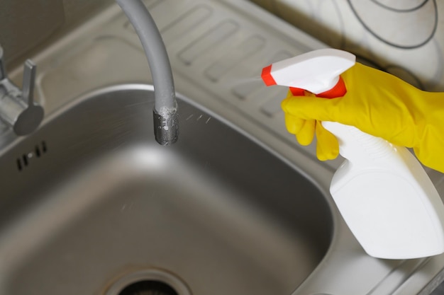 Una mujer con guantes de goma limpia el grifo y el fregadero en la cocina aplicando desinfectante de una botella de spray