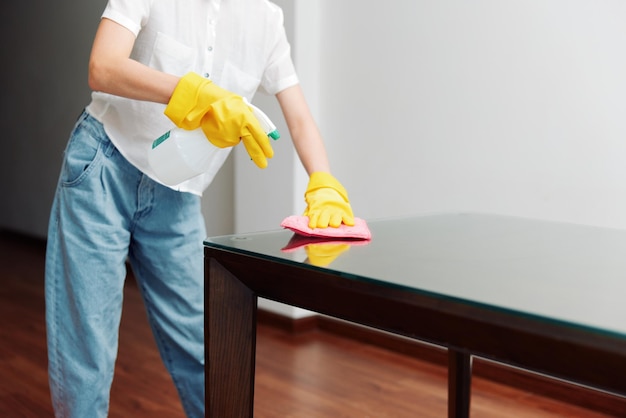 Mujer con guantes de goma limpiando una mesa de vidrio con una escoba y un cubo de agua en el fondo