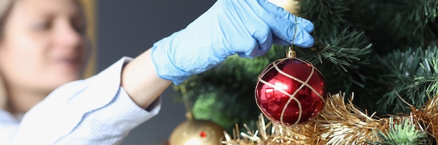 Mujer en guantes de goma protectores que cuelgan la bola en el primer del árbol de Navidad. Preparándose para el año nuevo durante la pandemia concepto covid19