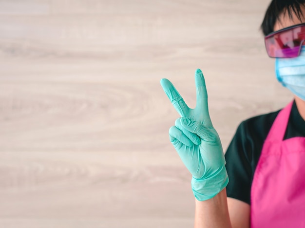 Mujer con guantes de látex y delantal rosa sosteniendo las manos como símbolo de victoria Ok, espero ganar médica