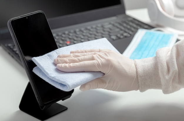 Mujer con guantes limpia phomelaptop con tejido húmedo y desinfectante durante el covid 19. Teléfono de desinfección y teclado de computadora portátil con desinfectante de alcohol por mujer con máscara brilla en el lugar de trabajo, escritorio de oficina.