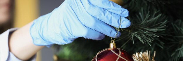 Mujer con guantes de protección cuelga una bola roja de año nuevo en las vacaciones del árbol de navidad por coronavirus