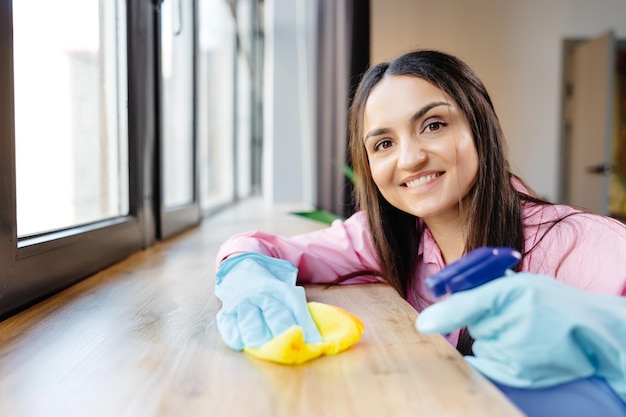 Una mujer con guantes protectores está sonriendo y limpiando el polvo con un pulverizador y un limpiador mientras limpia su casa de cerca