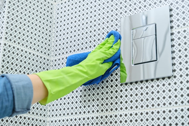 Mujer en guantes con trapo haciendo limpieza en el baño, limpiando y puliendo el botón de inodoro cromado en la pared de azulejos