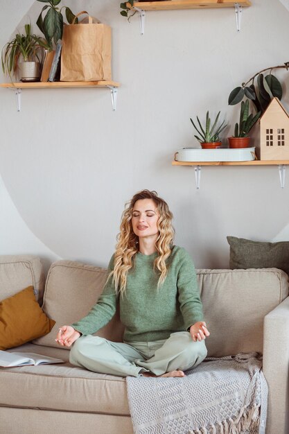 Una mujer guapa con un acogedor suéter verde sentada en el sofá y meditando en casa