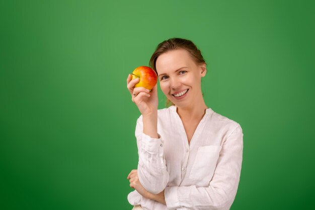 Una mujer guapa de cuarenta años con una camisa blanca sobre un fondo verde sonríe y sostiene una manzana fresca en sus manos Alimentación saludable vegetarianismo concepto de apariencia saludable