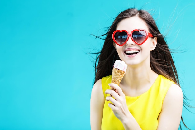 Mujer guapa morena vestida con camisa amarilla y gafas de sol comiendo helado de gofre antes del fondo azul