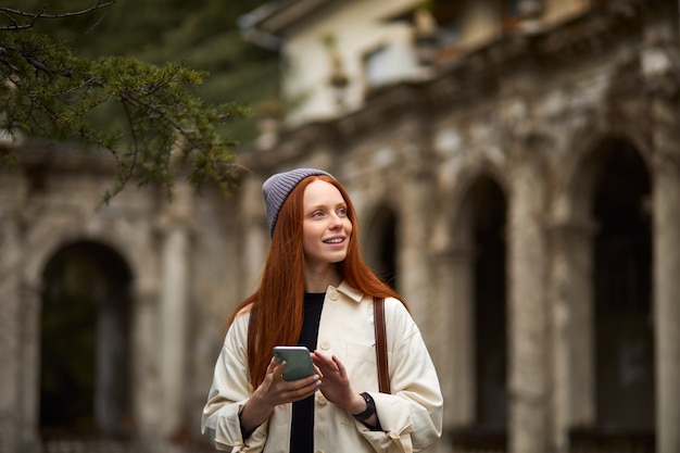 Mujer guapa de pie junto a un edificio antiguo con tecnología de pantalla táctil en el teléfono inteligente, navegar por Internet, mirar de lado, sonreír, pasar las vacaciones sola en otro país, caminar