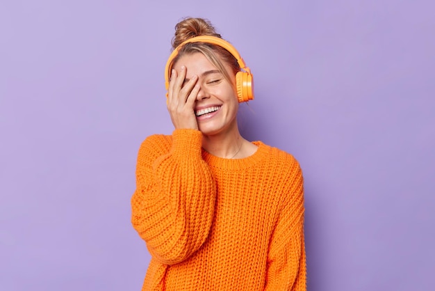 mujer hace cara palma sonríe feliz mantiene los ojos cerrados escucha la pista de audio a través de auriculares vestida con un jersey naranja de punto casual aislado en morado.