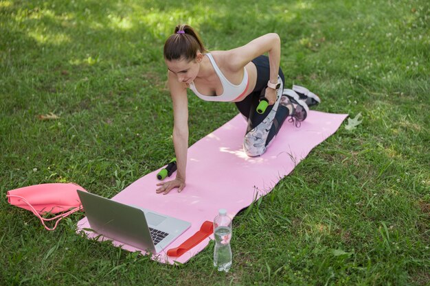 La mujer hace ejercicio con mancuernas viendo videos a través de una computadora portátil en el parque