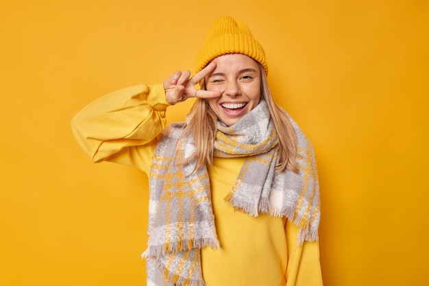 Foto la mujer hace un gesto de paz en los ojos, guiña un ojo y sonríe con alegría. el signo de la victoria lleva una bufanda informal alrededor del cuello y un sombrero aislado en amarillo. lenguaje corporal