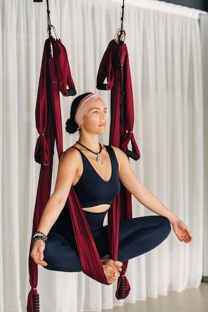 Una mujer hace yoga sentada en una hamaca colgante en el gimnasio.