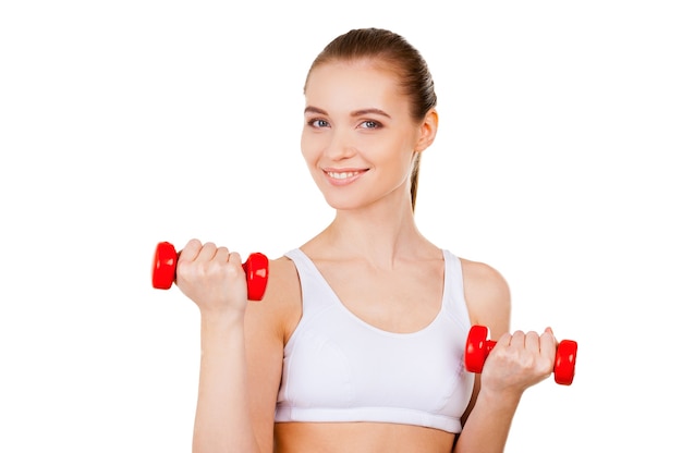 Mujer haciendo ejercicio con pesas. Atractiva mujer joven en ropa deportiva entrenando con pesas y sonriendo mientras está de pie aislado en blanco