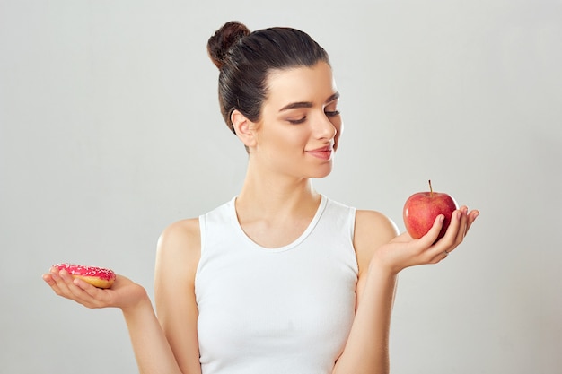 Mujer haciendo elección entre manzana y postre. Concepto de dieta. La muchacha morena sostiene una manzana y un postre rosado.