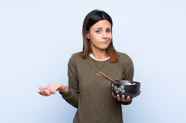 Mujer haciendo gesto de dudas mientras levanta los hombros mientras sostiene un tazón de fideos con palillos