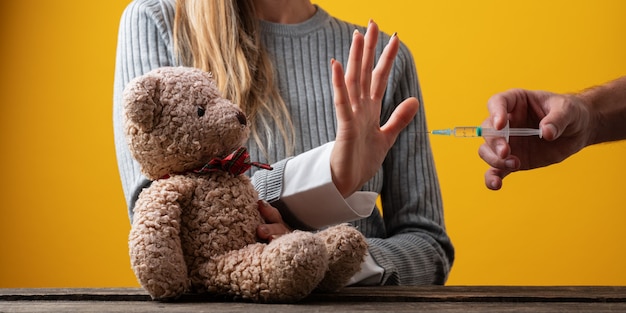 Mujer haciendo un gesto de parada con la mano hacia una vacuna mientras protege su osito de peluche en una imagen conceptual.