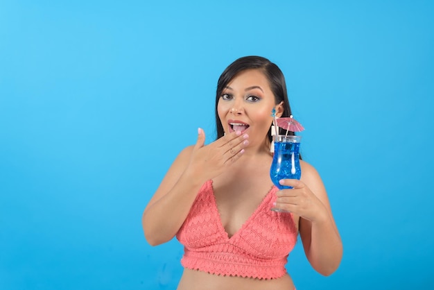 Mujer haciendo un gesto sorprendido sosteniendo un cóctel retrato de verano fondo azul