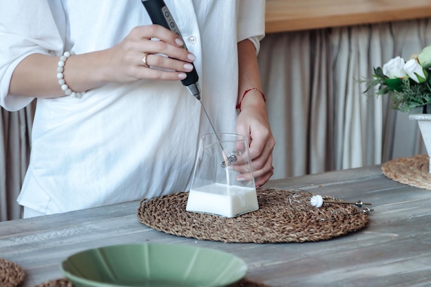 Mujer haciendo leche espumosa con batidora de mano mezclando leche en una  olla de vidrio con