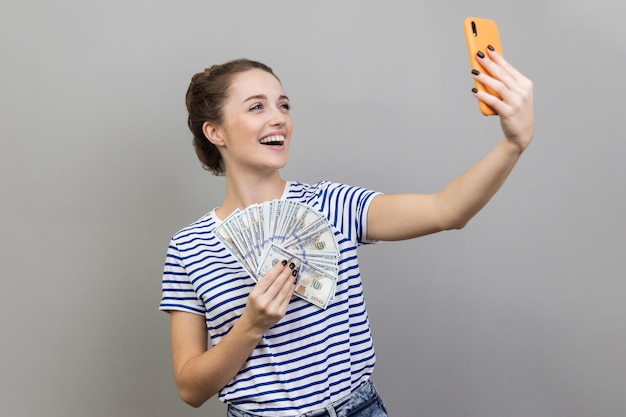 Mujer haciendo selfie con dinero escondiendo la cara detrás de un ventilador de billetes de cien dólares alardeando de ingresos