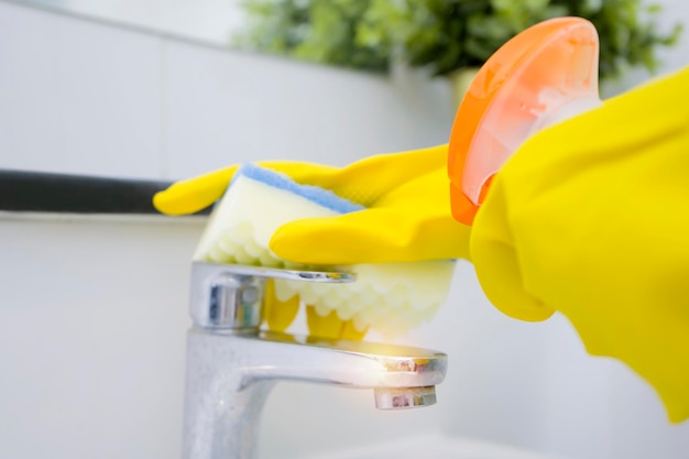 Mujer haciendo tareas en el baño en casa limpiando el fregadero y el grifo con detergente en aerosol Vista recortada Mano de sirvienta o mujer limpiando un nuevo lavabo moderno limpio con guantes amarillos y una esponja