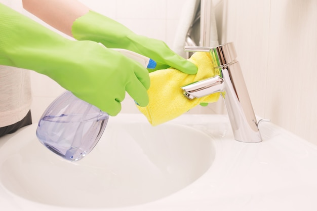 Mujer haciendo tareas domésticas en el baño, limpiando el fregadero y el grifo con spray.