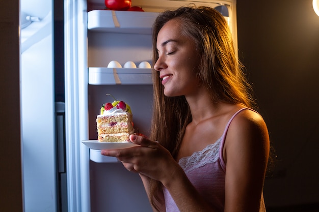 Mujer hambrienta en pijama come pastel dulce por la noche cerca del refrigerador. Deje de hacer dieta y aumente de peso debido a los carbohidratos y la alimentación poco saludable.