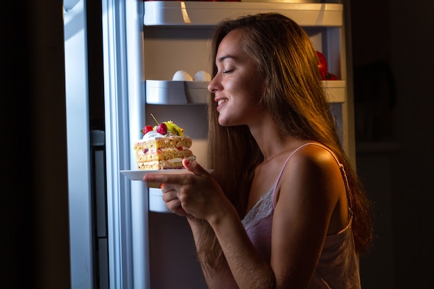 Mujer hambrienta en pijama comiendo pastel dulce en la noche cerca de nevera