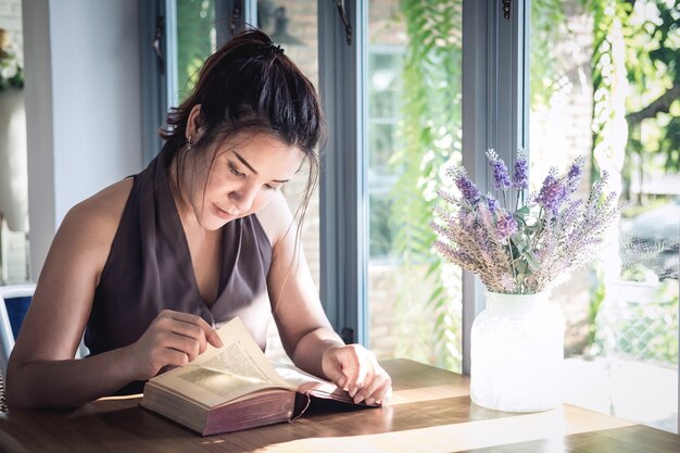 Mujer hermosa asiática joven que se sienta en la tabla y que lee el libro interesante