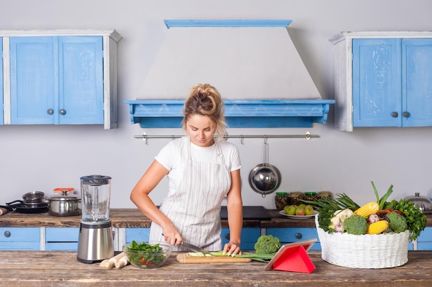 Una mujer hermosa en delantal cortando verduras cocinando ensalada en la cocina preparando comida vegetariana siguiendo la receta en internet viendo una cesta de tabletas de verduras verdes frescas en la mesa