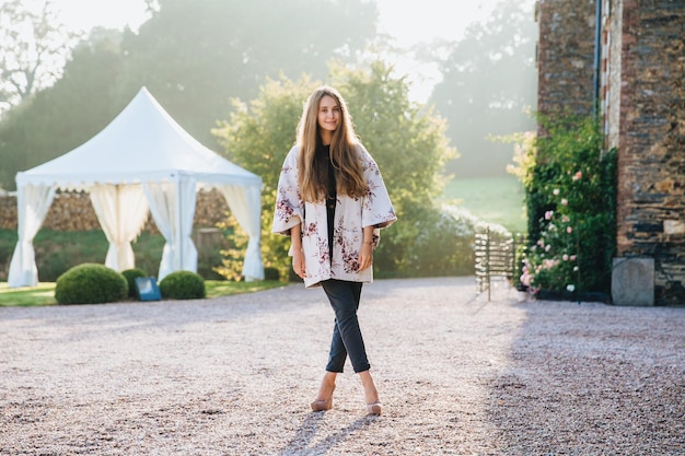 Una mujer hermosa y elegante viste ropa de moda, tiene el pelo largo y liso, las piernas cruzadas contra la hermosa naturaleza, un edificio antiguo y una carpa blanca, disfruta del sol, tiene una excursión en un lugar desconocido