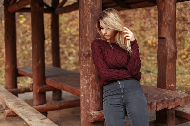 Mujer hermosa con estilo en un suéter de moda con jeans posando cerca de una glorieta de madera