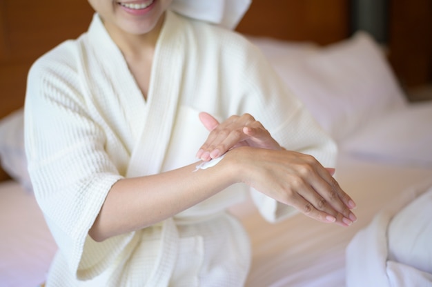 Una mujer hermosa feliz en bata de baño blanca aplicando crema hidratante corporal en el concepto de dormitorio, cuidado de la piel y tratamiento