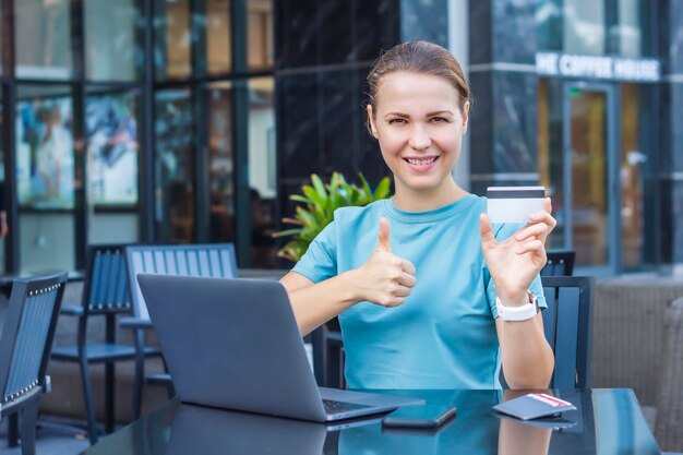 La mujer hermosa feliz que disfruta de compras en línea que sonríe con la computadora portátil, teléfono inteligente que sostiene la tarjeta de crédito en la mano que se sienta en café en terraza al aire libre, muestra el pulgar para arriba, como gesto. Concepto de pago en línea