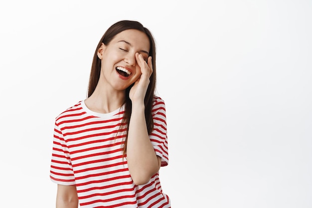 Mujer hermosa feliz riendo, tocando la cara con los ojos cerrados y la expresión tierna de la cara, sonriendo despreocupada, parada en camiseta contra fondo blanco