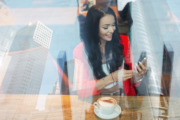 Mujer hermosa feliz con una sonrisa en ropa de moda está sentada en un café tomando café y charlando en el teléfono inteligente Chica guapa con emoción detrás del cristal con un reflejo de la ciudad