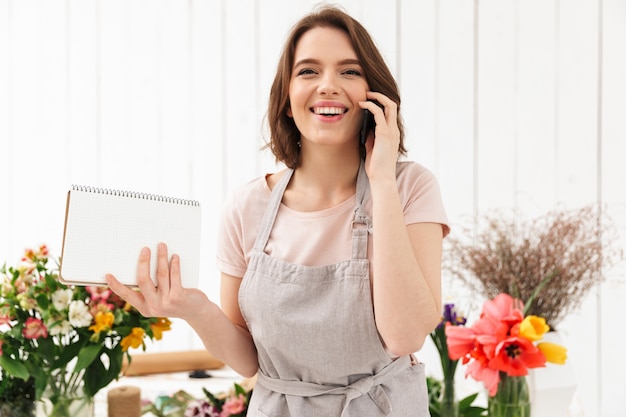 Mujer hermosa floristería en delantal trabajando en flor y hablando por teléfono móvil con notas en la mano