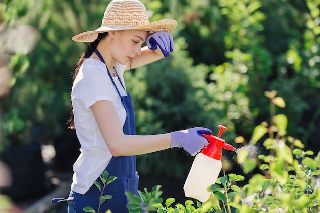 Mujer hermosa jardinera con sombrero de paja rocía plantas de un rociador de jardín.